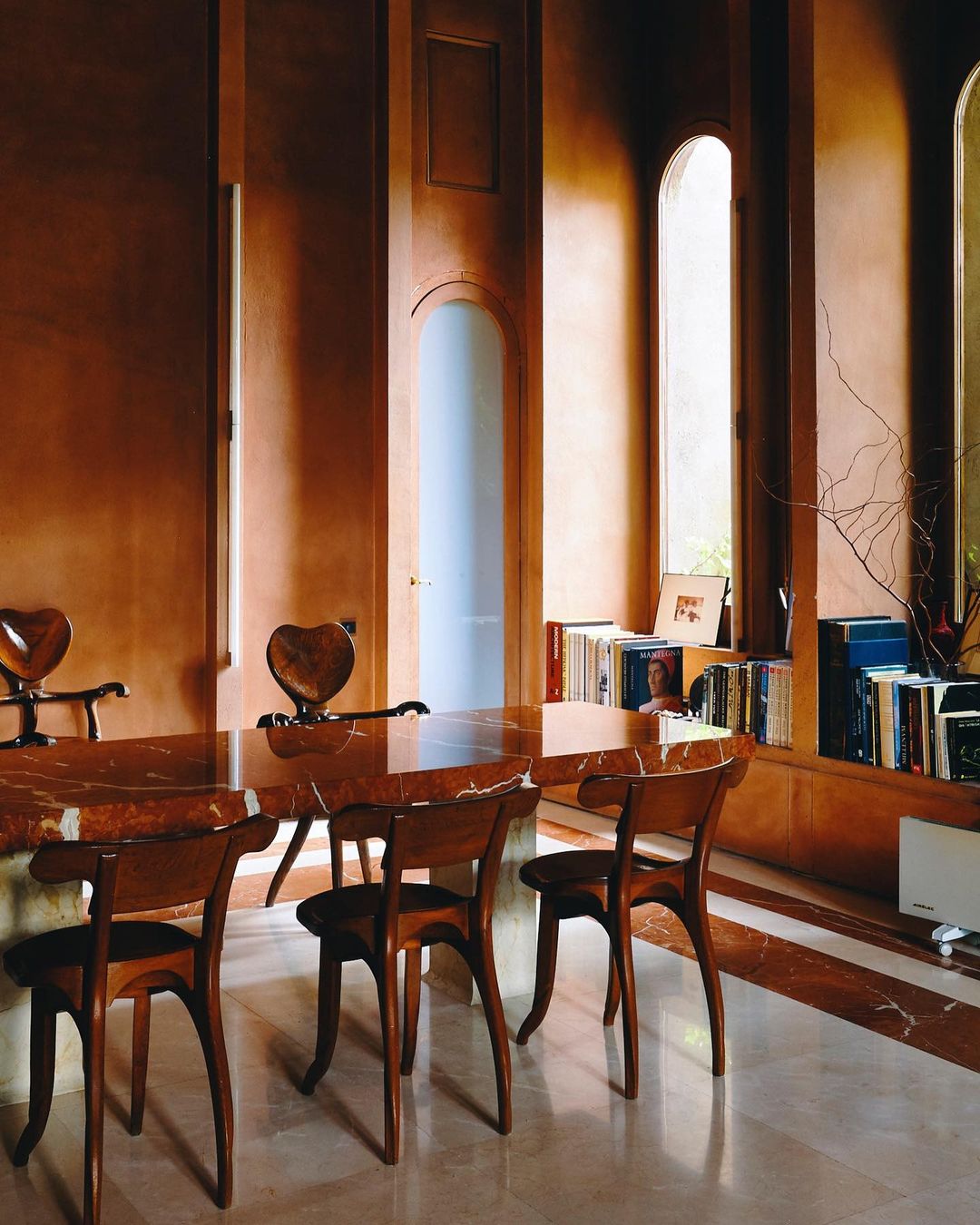 Interior view of a dining room featuring a marble table and wooden chairs, with natural light filtering through arched windows.