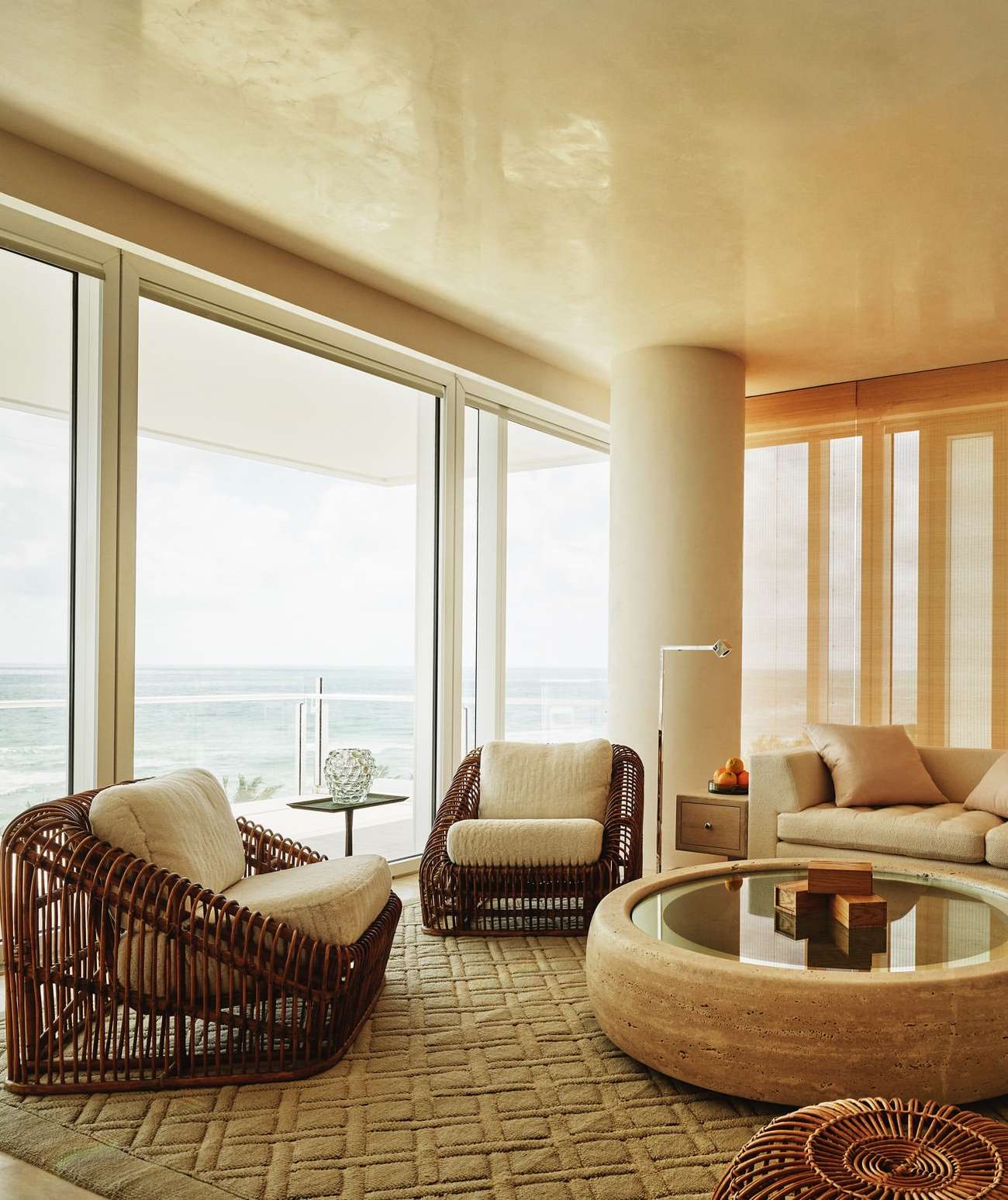 Living room interior featuring two brown wicker armchairs, a travertine table, a beige sofa, and a view of the ocean through large windows.