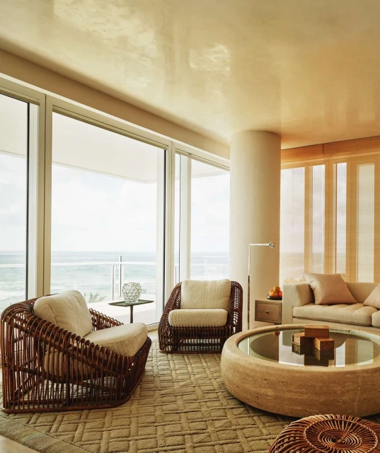Living room interior featuring two brown wicker armchairs, a travertine table, a beige sofa, and a view of the ocean through large windows.