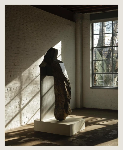 Abstract stone sculpture with metal element stands on a white base inside a brick-walled room, illuminated by window light and shadows.
