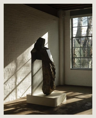 Abstract stone sculpture with metal element stands on a white base inside a brick-walled room, illuminated by window light and shadows.
