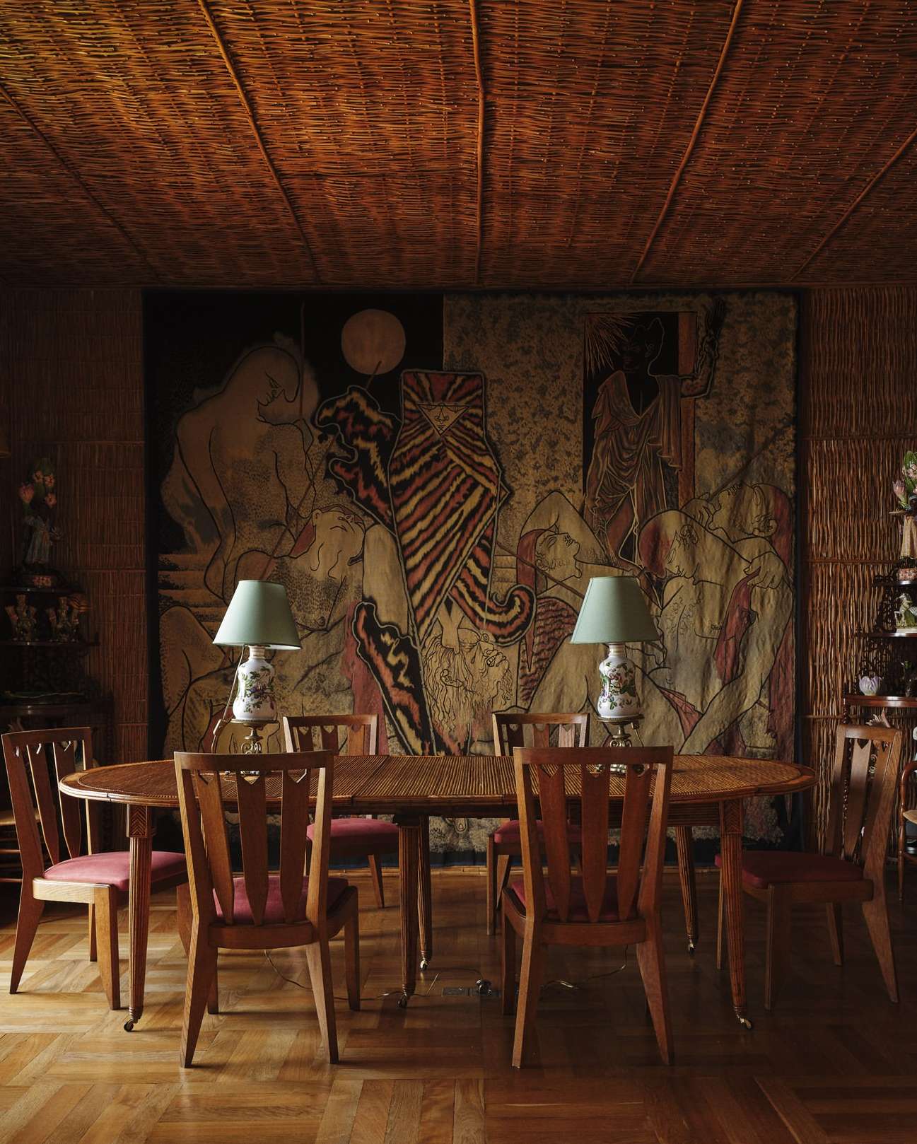 Interior view of a dining room with a wooden table, chairs, patterned tapestry, woven ceiling, and parquet flooring; a warm and inviting setting.