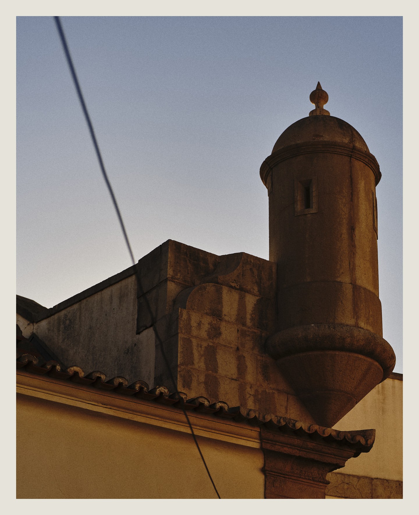 Low-angle shot of a historic stone tower with a domed top, integrated into a building with a tiled roof under a clear, light-blue sky.
