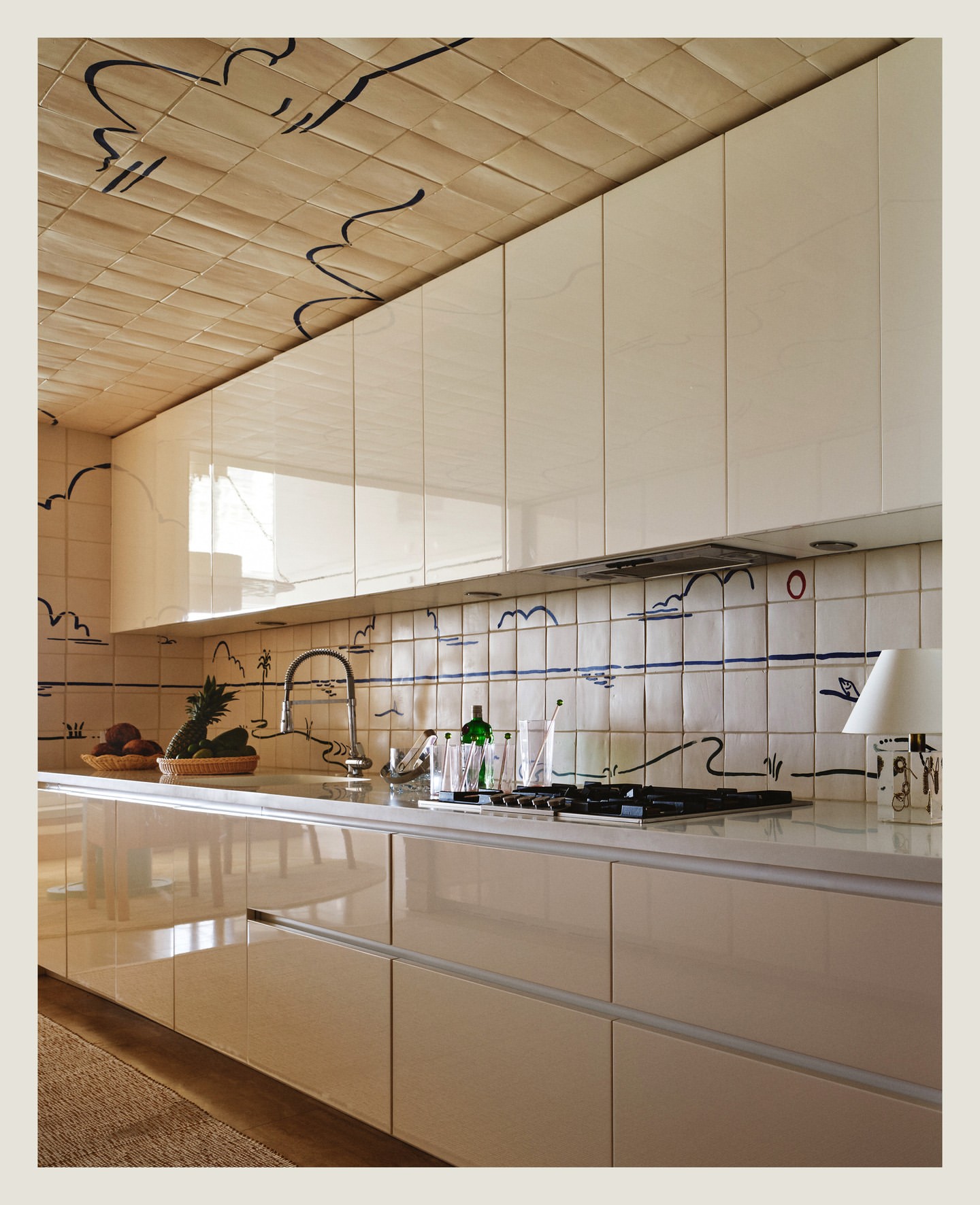 Interior shot of a modern kitchen featuring light-colored cabinetry, a countertop with fruits and glasses, and decorative tile work.