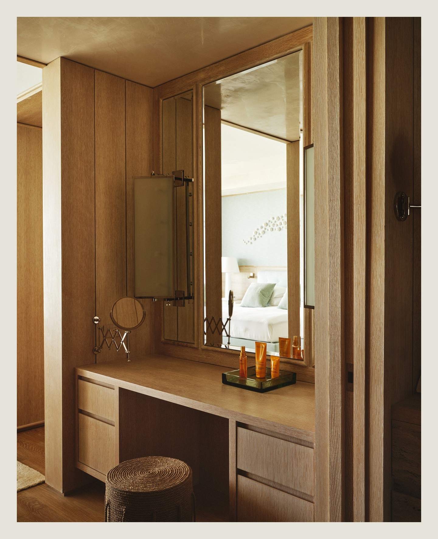 A wooden vanity with a mirror in a bedroom featuring a stool, cosmetic products, and a glimpse of a bed with light-colored linens.