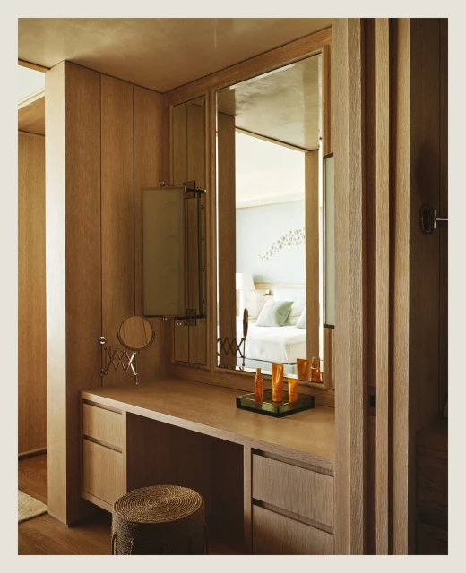 A wooden vanity with a mirror in a bedroom featuring a stool, cosmetic products, and a glimpse of a bed with light-colored linens.