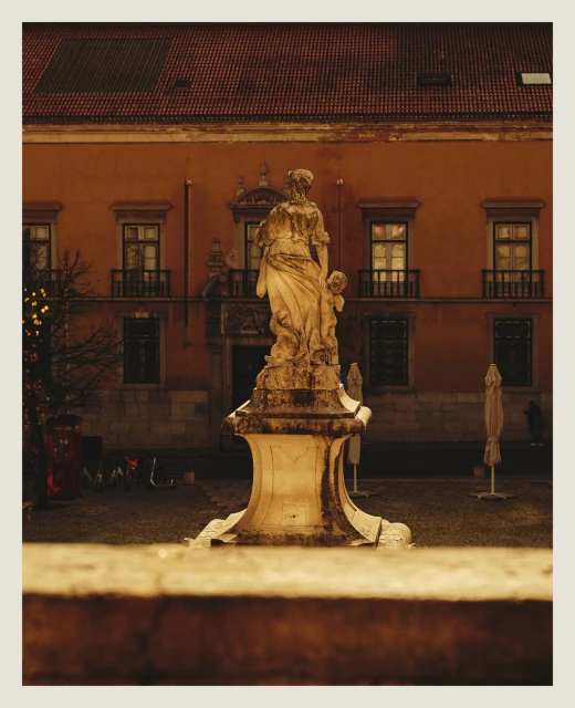 Outdoor photo of a marble sculpture featuring two figures in a courtyard with an orange building facade and windows in the background.