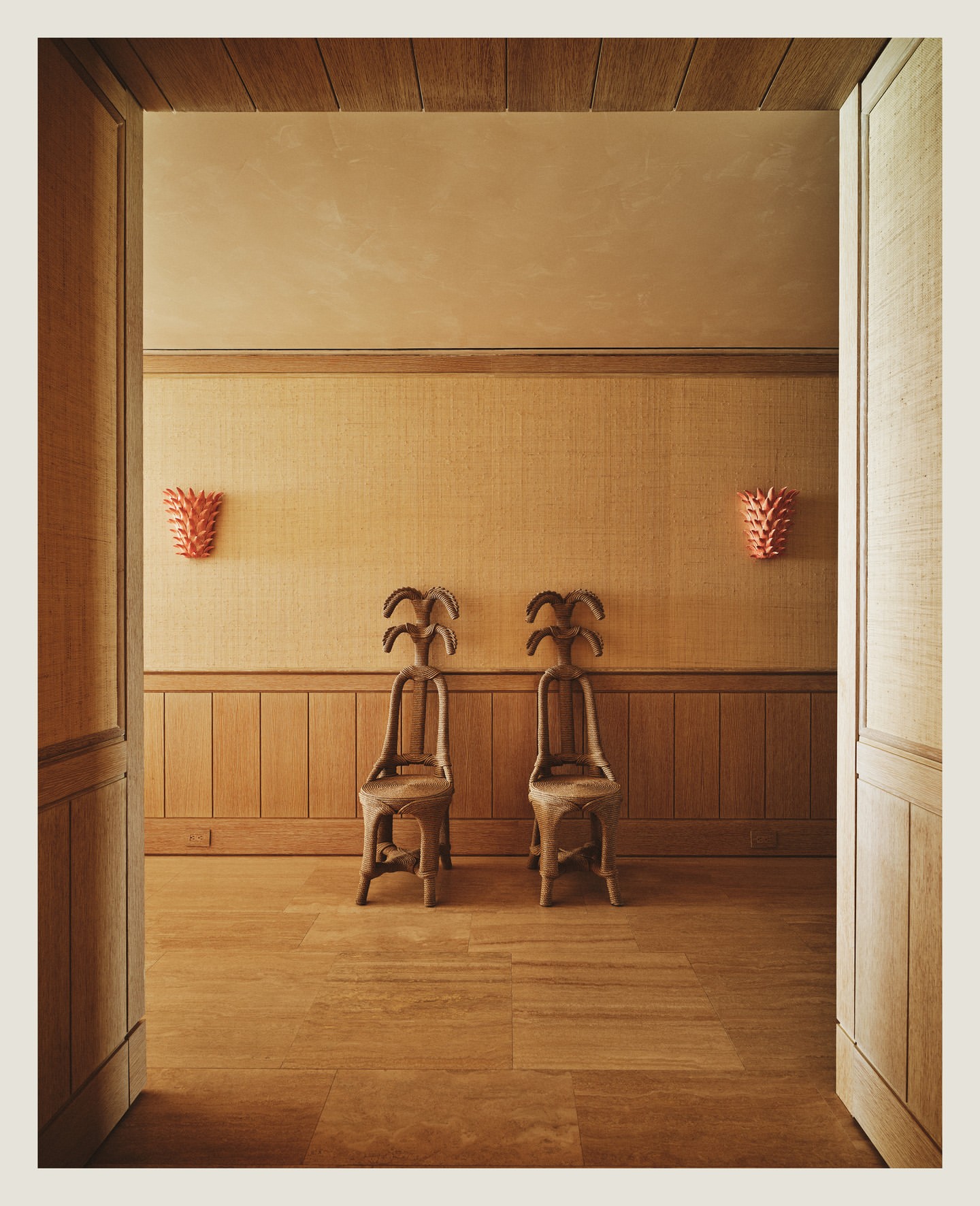 Interior design with two ornate wooden chairs on a wooden floor, coral sconces on the wall, and an open doorway, creating an inviting space.