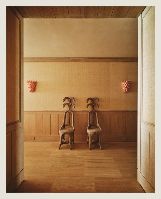Interior design with two ornate wooden chairs on a wooden floor, coral sconces on the wall, and an open doorway, creating an inviting space.