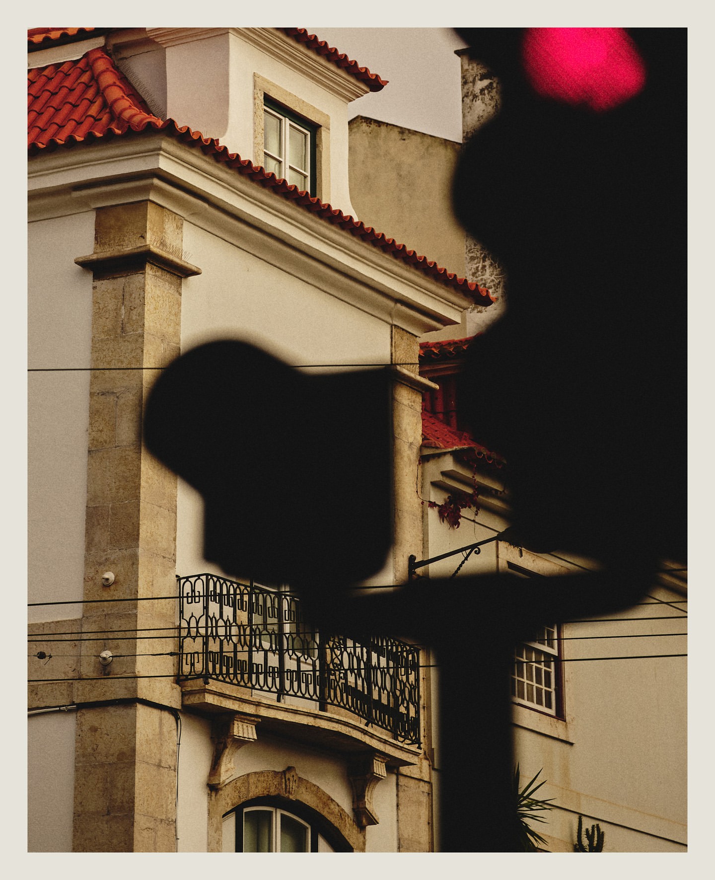Exterior architectural image of a cream colored building with a red tile roof, black wrought iron balcony, and dark obscured foreground.