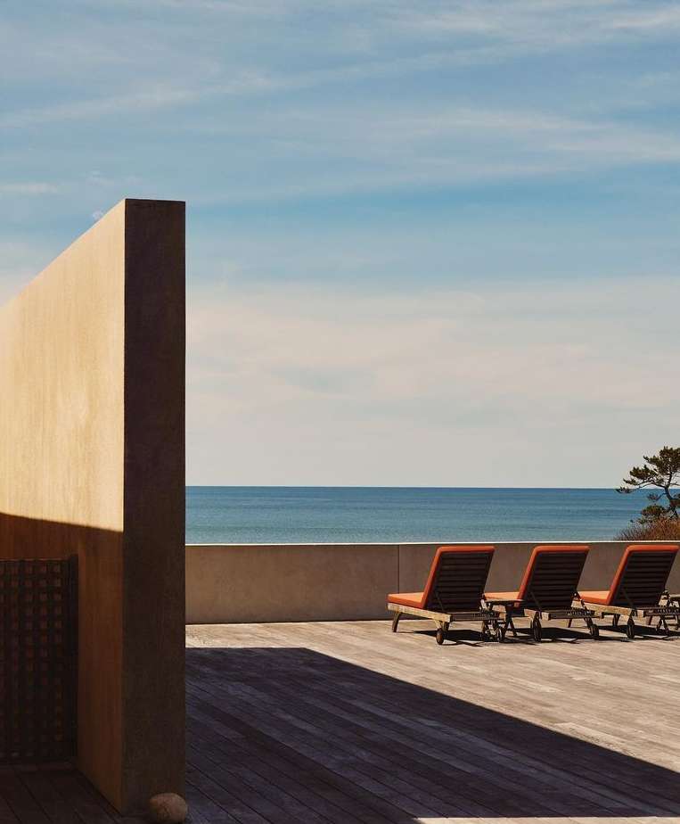 Outdoor patio with three lounge chairs on a wooden deck overlooking the ocean under a clear blue sky, next to a beige wall.