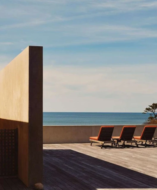 Outdoor patio with three lounge chairs on a wooden deck overlooking the ocean under a clear blue sky, next to a beige wall.