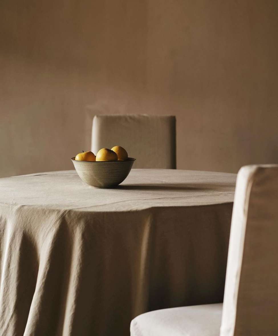 Still life composition with a bowl of lemons on a table with linen, set in an indoor room with soft, diffused lighting.
