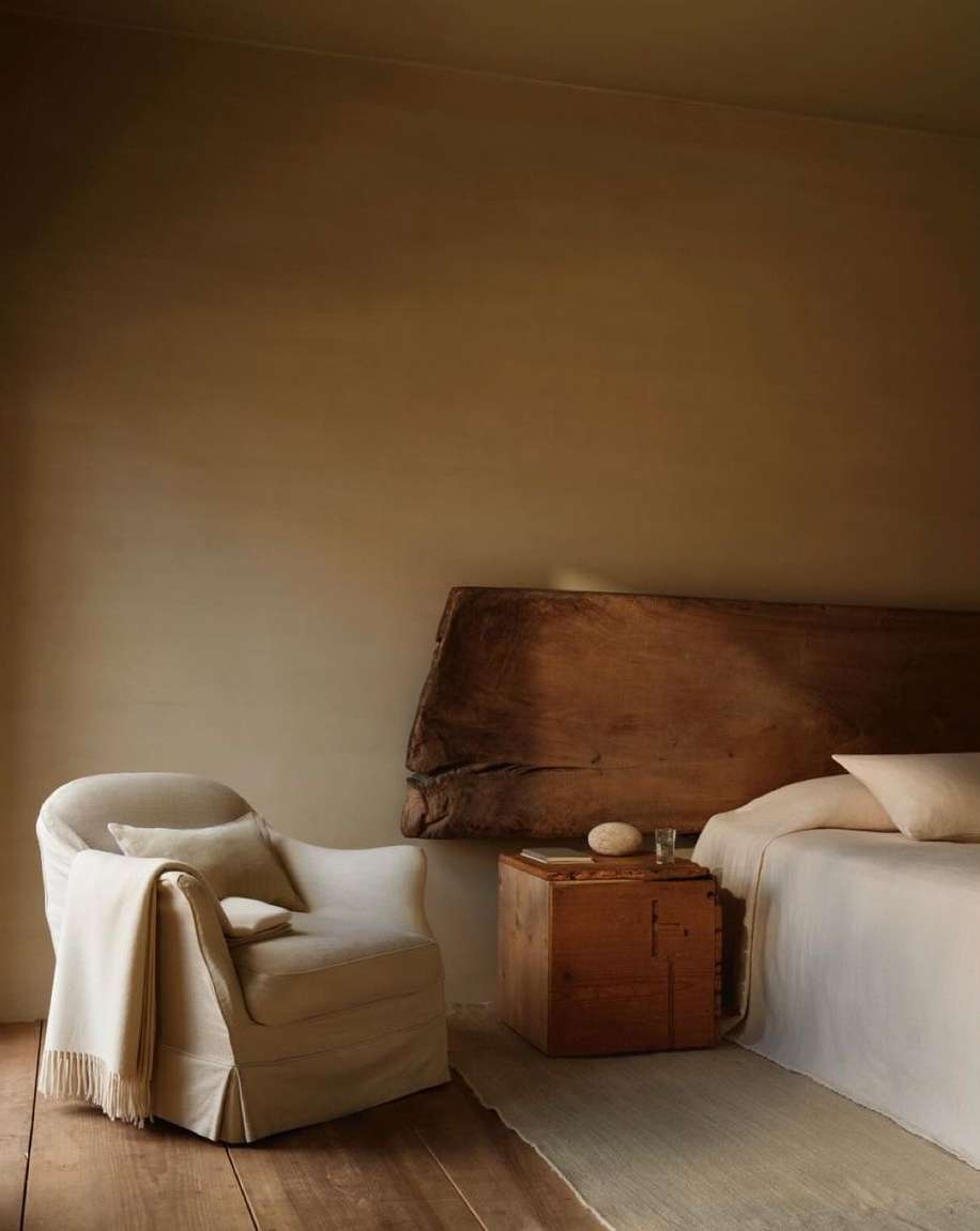 Interior with an armchair draped in a blanket, a wooden side table, and a bed with white linens set against a neutral wall.