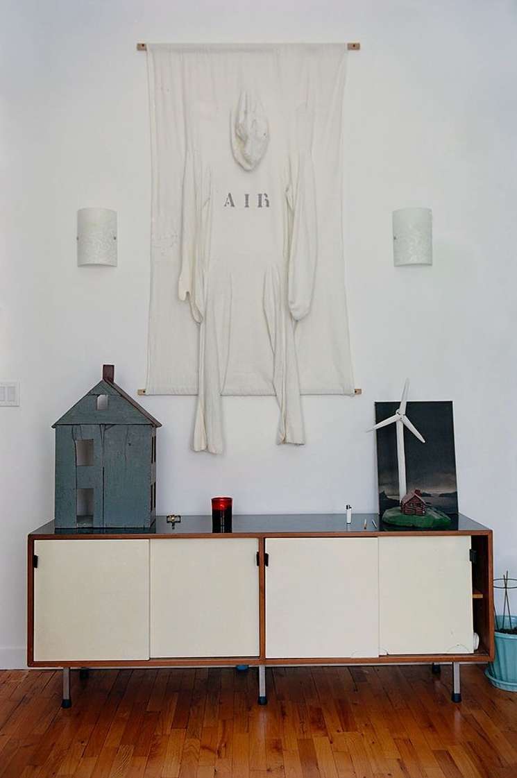 Art installation featuring a white jumpsuit hanging against a white wall, displayed above a mid-century modern credenza, and a model house.