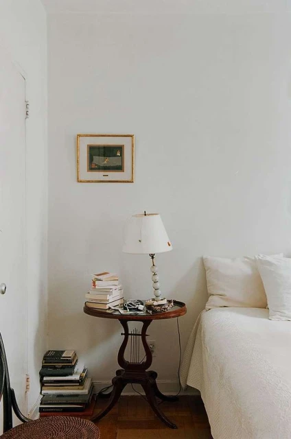Bedroom interior with a small wooden table holding books and a lamp next to a bed, with a neutral color palette and a cozy, inviting atmosphere.