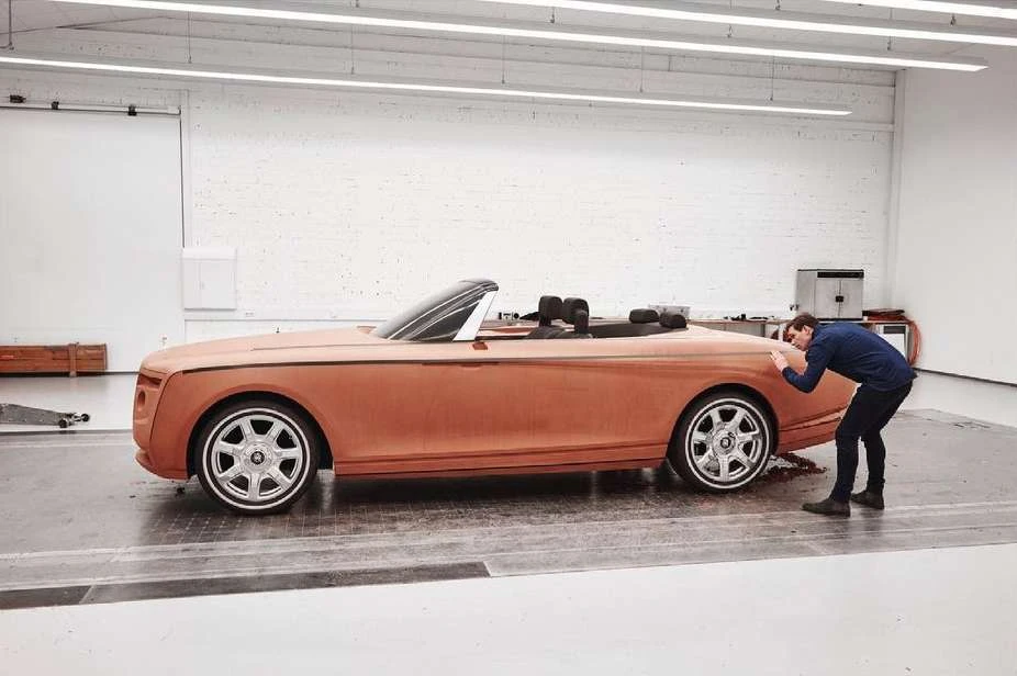 Unfinished clay-colored convertible car is worked on by an adult male in an indoor studio, with a minimalist white brick background.