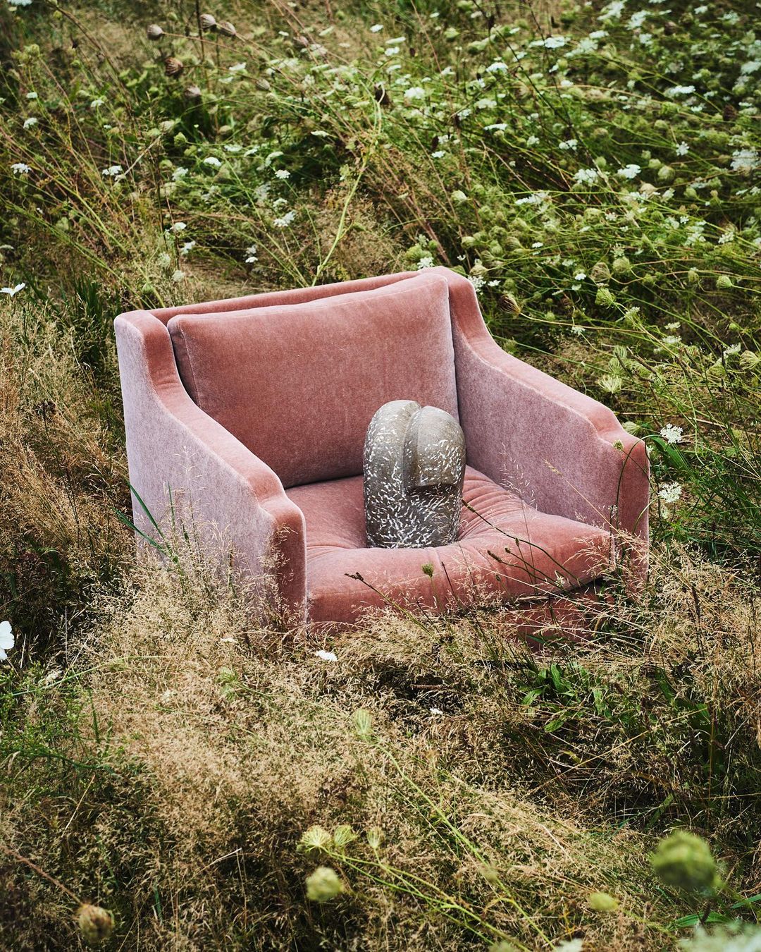Pink armchair sits amidst tall grass and wildflowers, creating a surreal blend of domestic comfort and natural wilderness in a quiet outdoor setting.