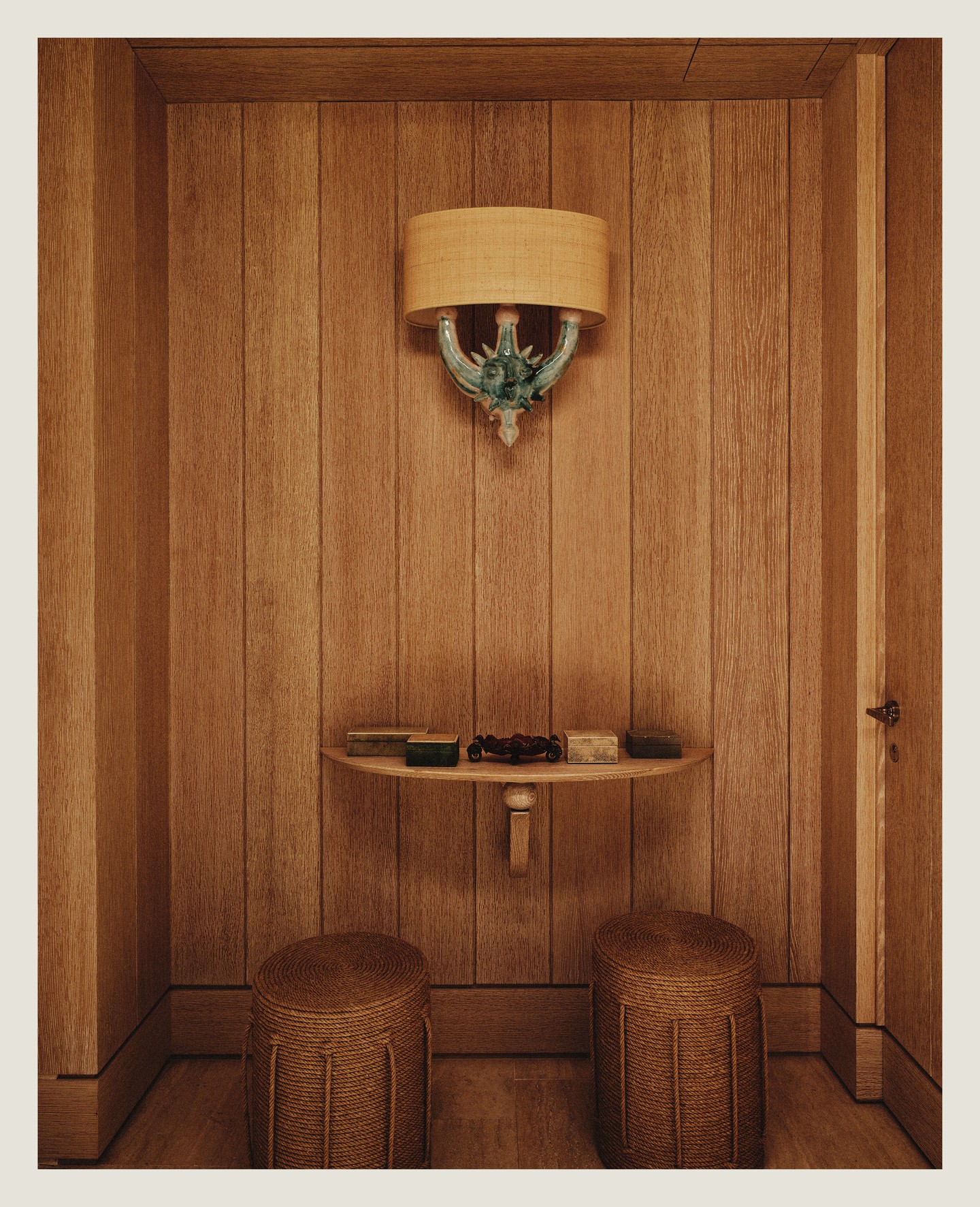 Interior shot of a wood-paneled room with a light fixture, small table holding various small boxes, and two wicker stools.