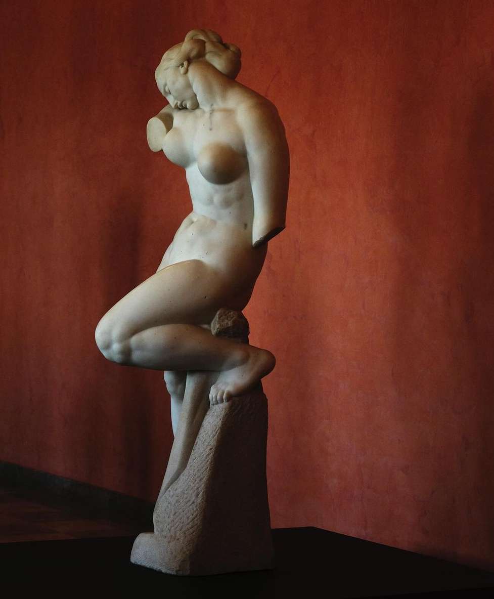 A nude marble statue of a woman stands indoors, illuminated by a spotlight against a terracotta colored wall in an art setting.