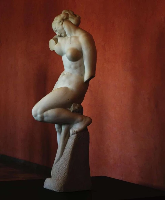 A nude marble statue of a woman stands indoors, illuminated by a spotlight against a terracotta colored wall in an art setting.