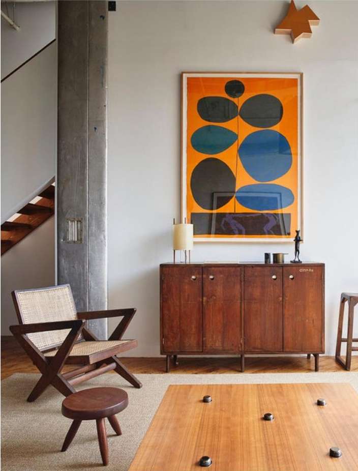 Living room interior with a wooden armchair, a round stool, a wooden sideboard cabinet, and a modern art print on the wall.