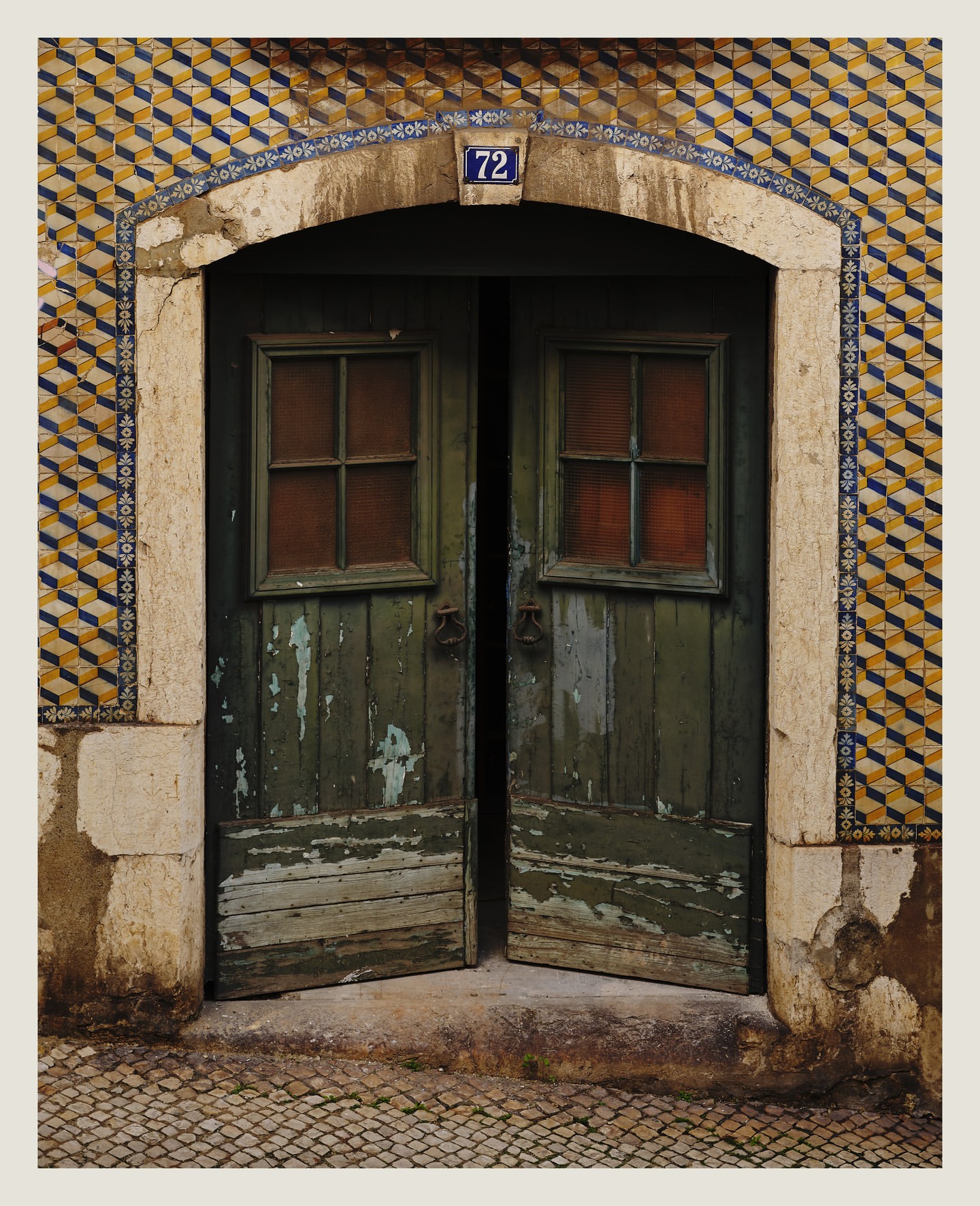 Weathered green wooden doors with small windows open to a stone-paved street, set within a tiled archway with the number 72 visible.