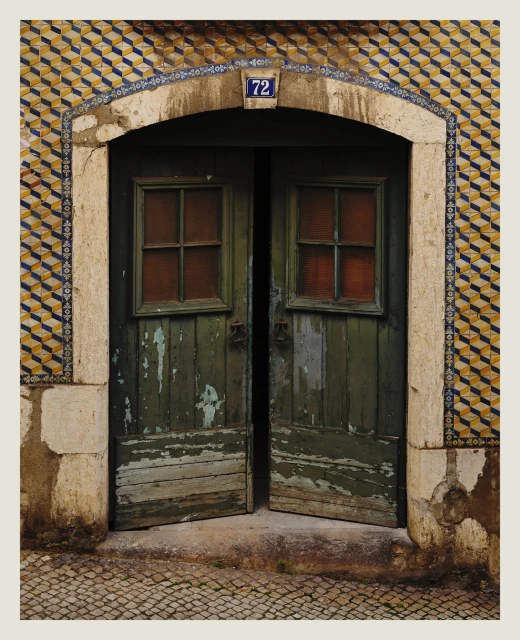 Weathered green wooden doors with small windows open to a stone-paved street, set within a tiled archway with the number 72 visible.