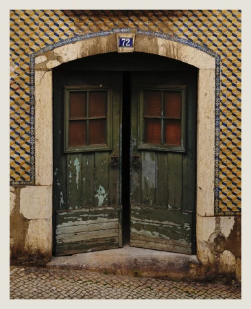 Weathered green wooden doors with small windows open to a stone-paved street, set within a tiled archway with the number 72 visible.