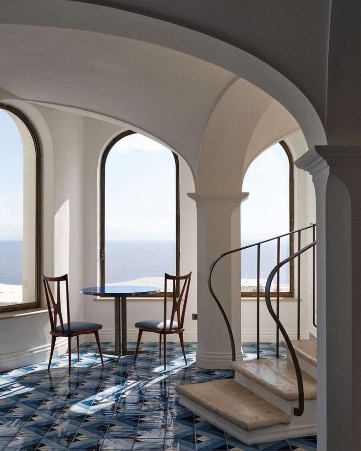 Interior view of a sunroom featuring arched windows offering a sea view, with a round table and two chairs on a blue patterned tile floor.