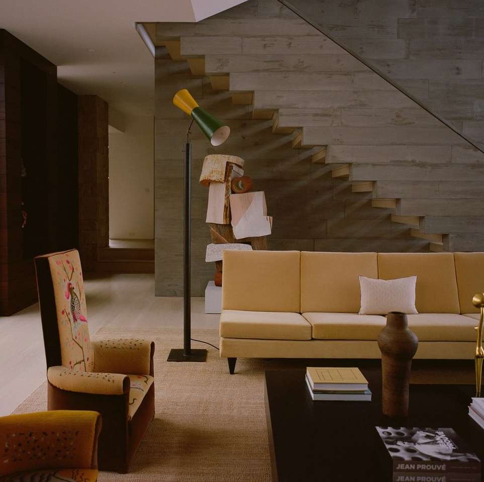 Interior of living room featuring a tan sofa, patterned armchairs, contemporary floor lamp, area rug, and concrete staircase in background.