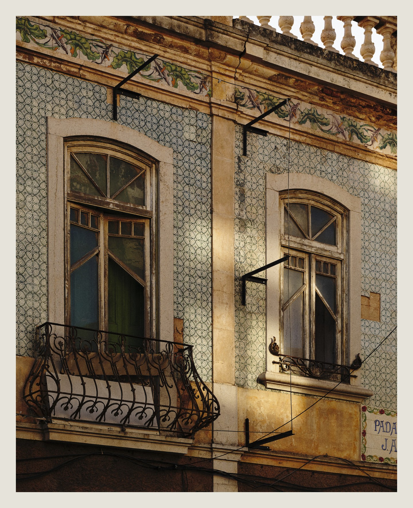 Close-up of a building facade with patterned tiles, featuring two windows, one with a wrought iron balcony, in soft, natural light.