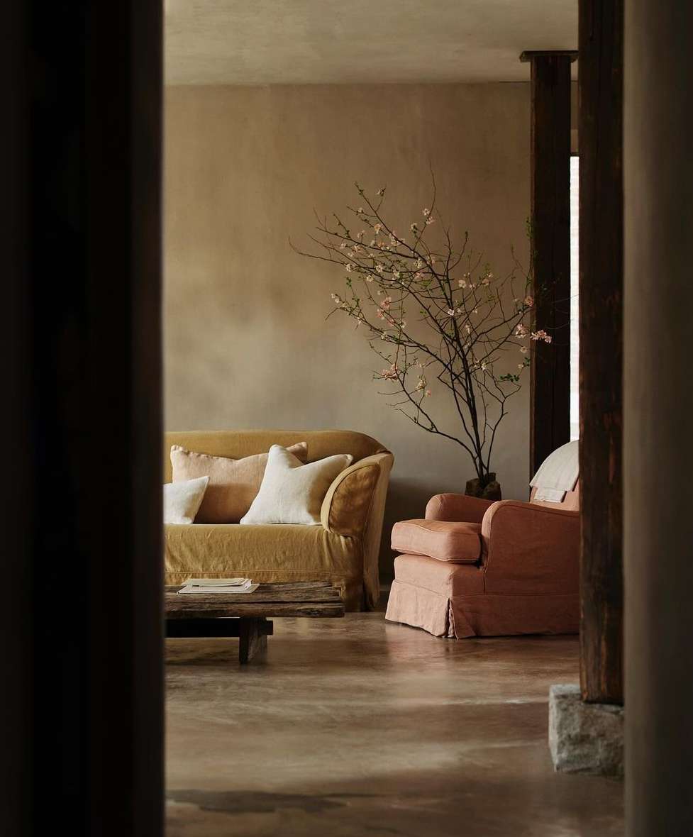 Living room with a yellow sofa, pink chair, and a bare tree branch in a vase set against a beige wall, creating a cozy interior.