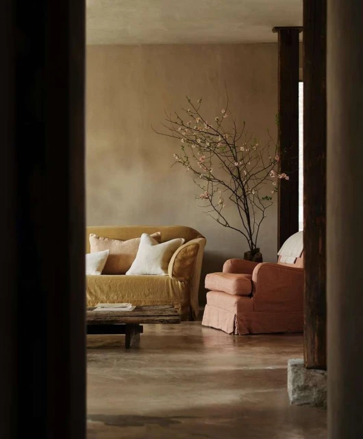 Living room with a yellow sofa, pink chair, and a bare tree branch in a vase set against a beige wall, creating a cozy interior.