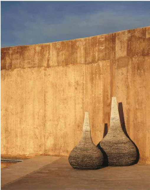 Two textured vases stand outdoors against a tall tan wall beneath a blue sky, exhibiting shades of brown, tan, and blue.