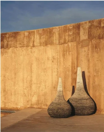 Two textured vases stand outdoors against a tall tan wall beneath a blue sky, exhibiting shades of brown, tan, and blue.