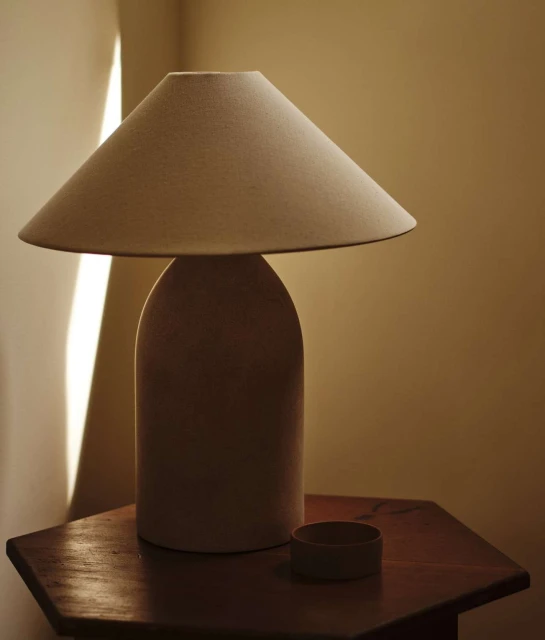 Beige table lamp on an octagonal table against a wall, illuminated by natural light, casting soft shadows in an indoor setting.