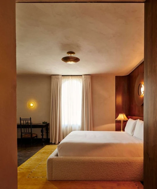 Interior view of a bedroom featuring a bed with white linens, a yellow rug, wooden desk, and warm lighting from lamps and sconces.