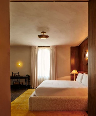 Interior view of a bedroom featuring a bed with white linens, a yellow rug, wooden desk, and warm lighting from lamps and sconces.