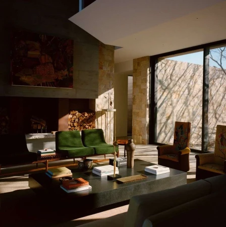 Living room interior with stone walls, green chairs, large dark coffee table, books, and a fireplace with stacked wood logs.