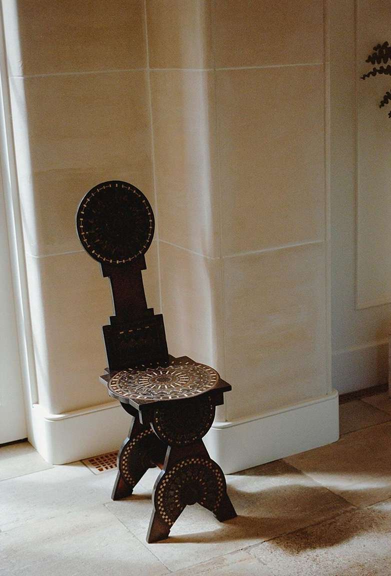 Dark wood antique chair with inlaid patterns and decorative circles near white wall with sunlight and shadow in interior setting.