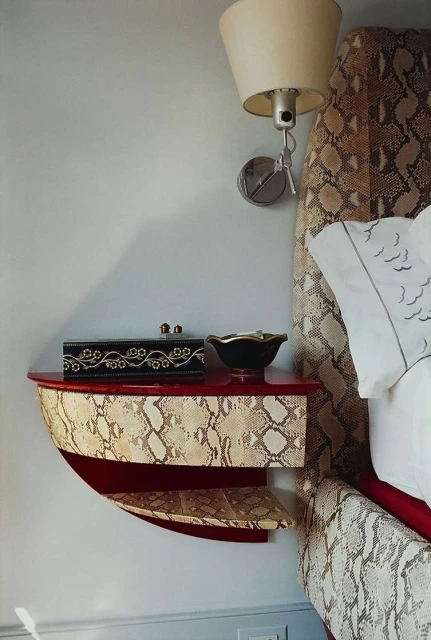 Snakeskin pattern headboard and red nightstand shelf with decorative black box and bowl in a bedroom interior, lit by a sconce.