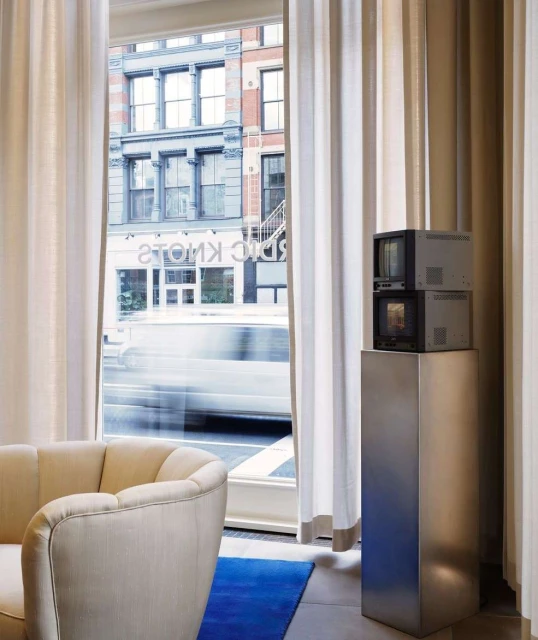 Living room interior featuring a modern beige sofa, a cobalt rug, two vintage television sets, and neutral-toned curtains.