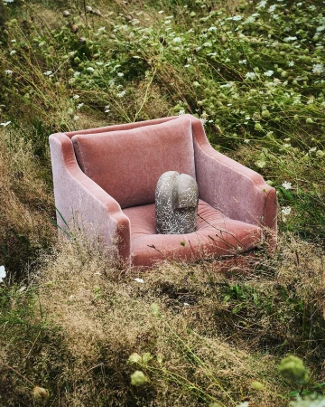 A pink velvet armchair sits outdoors nestled in a lush green meadow, holding a grey textured sculptural object on its seat cushion.
