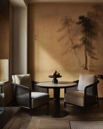A round wooden table with a bowl of fruit and two armchairs set against a textured wall with a tree mural.