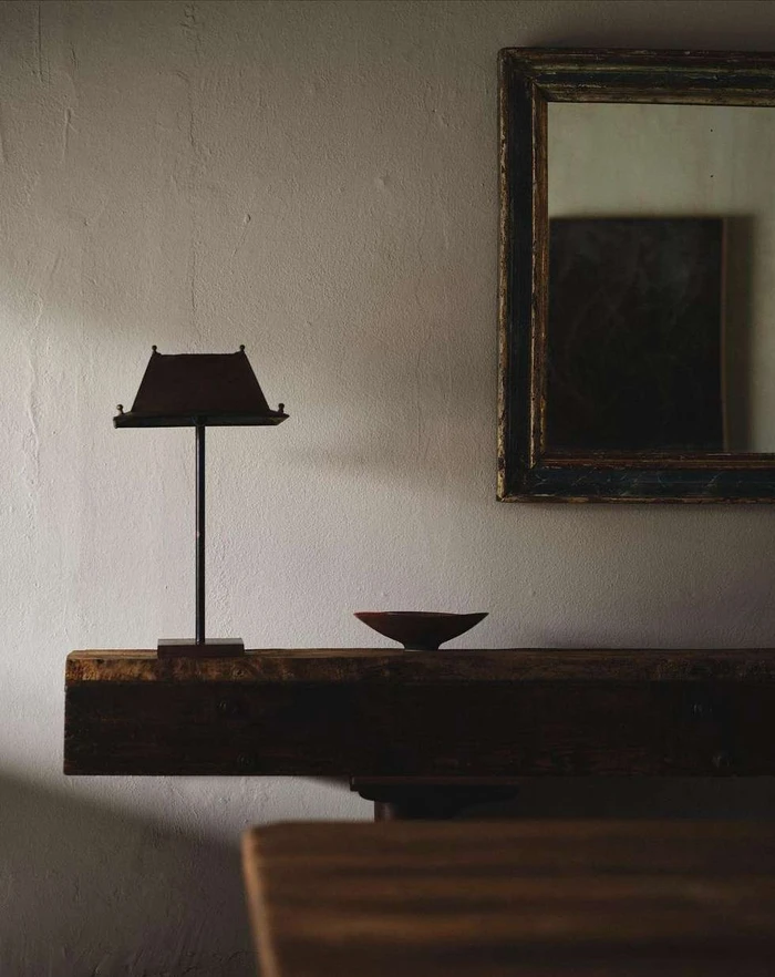 Minimalist interior with a wooden console table, a dark lamp, a small bowl, and a framed mirror on a textured wall.