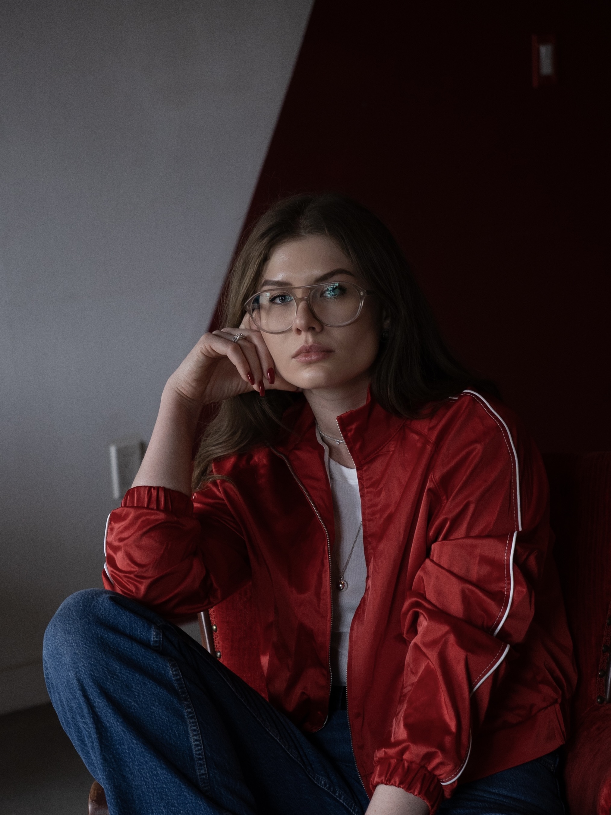 Portrait of a young woman with glasses, wearing a red jacket and jeans, seated indoors against a two-toned wall.
