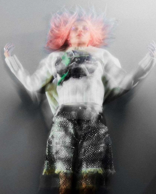 Woman with pink hair poses in a patterned sweater and sequined shorts against a gray background with a blurred motion effect.