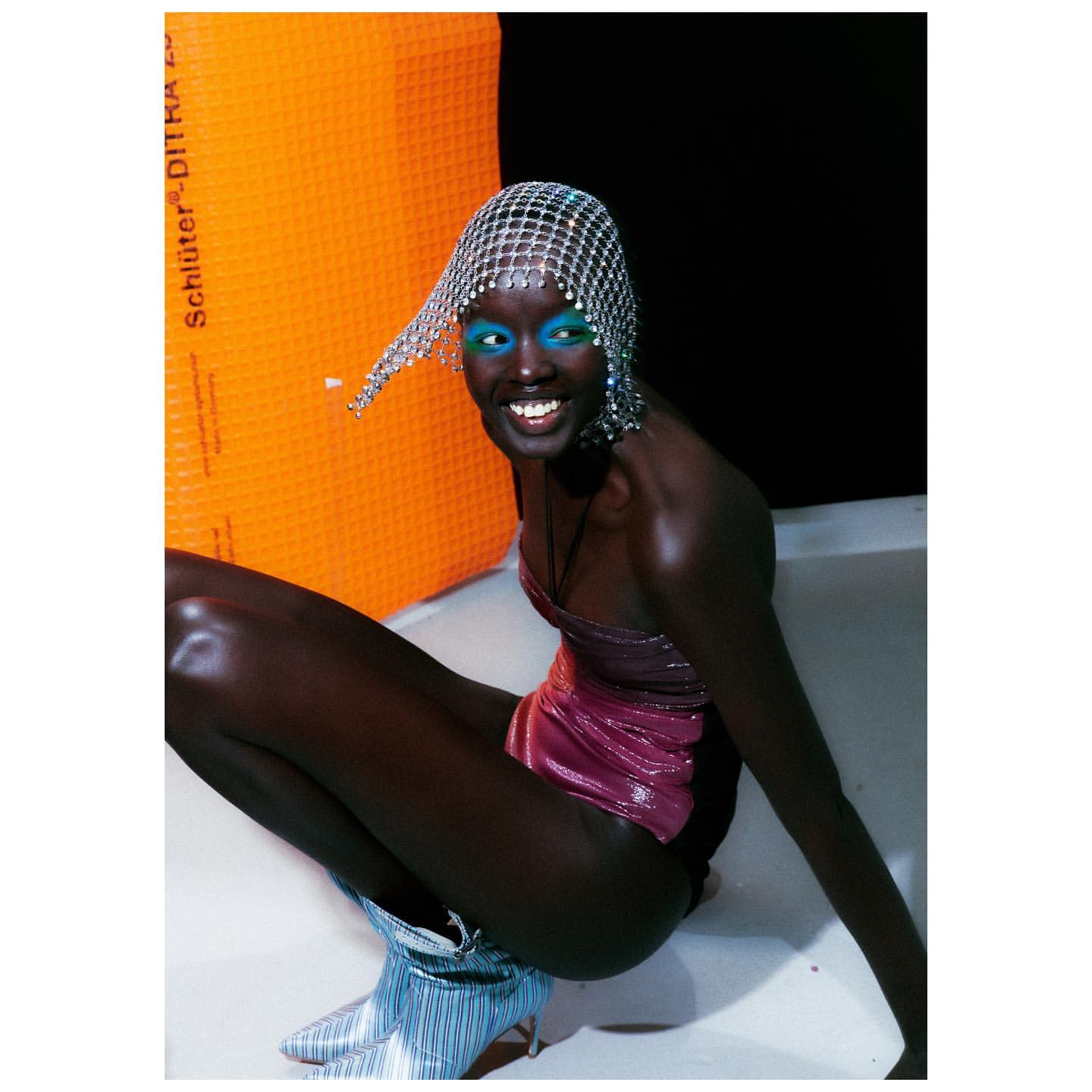 Smiling African woman poses in a shiny pink bodysuit, silver boots, and a mesh head covering against a dark and orange background.