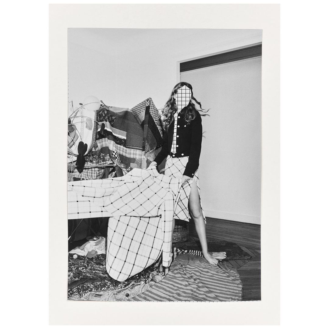 Monochrome portrait of a woman wearing a plaid skirt, jacket, and tie, posed indoors with clothing racks and patterned fabrics.